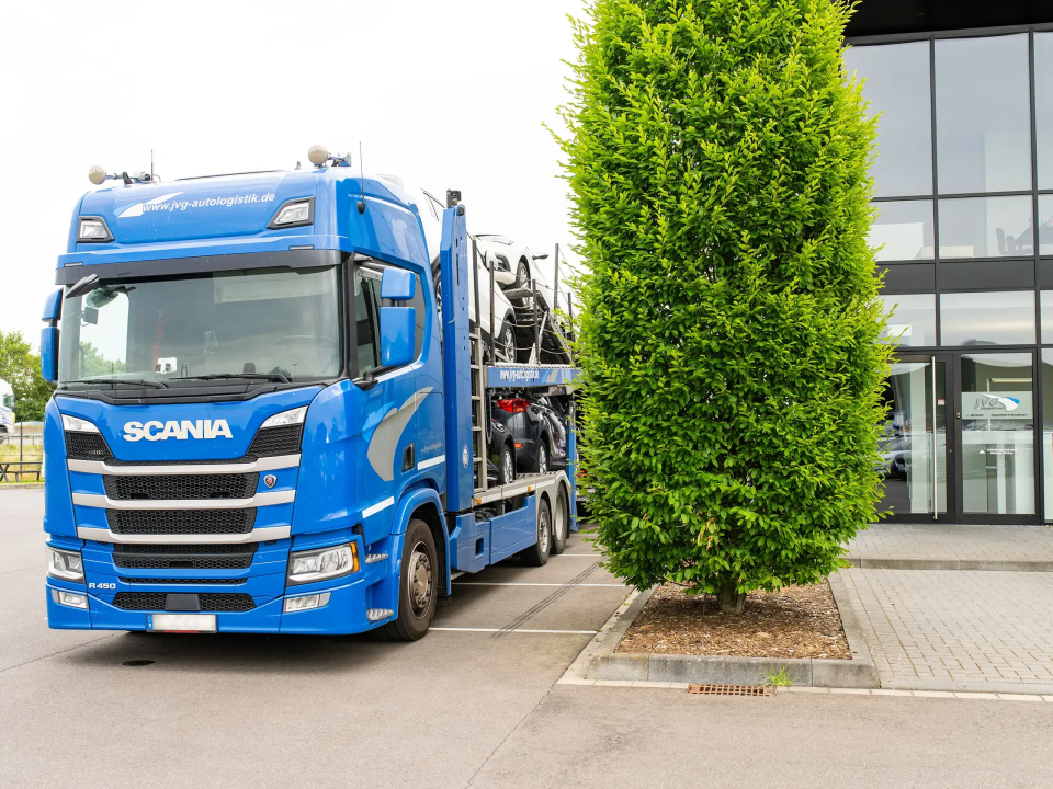 Blauer Scania LKW mit Auflieger steht vor dem JVG Firmengebäude, rechts ein großer grüner Baum.