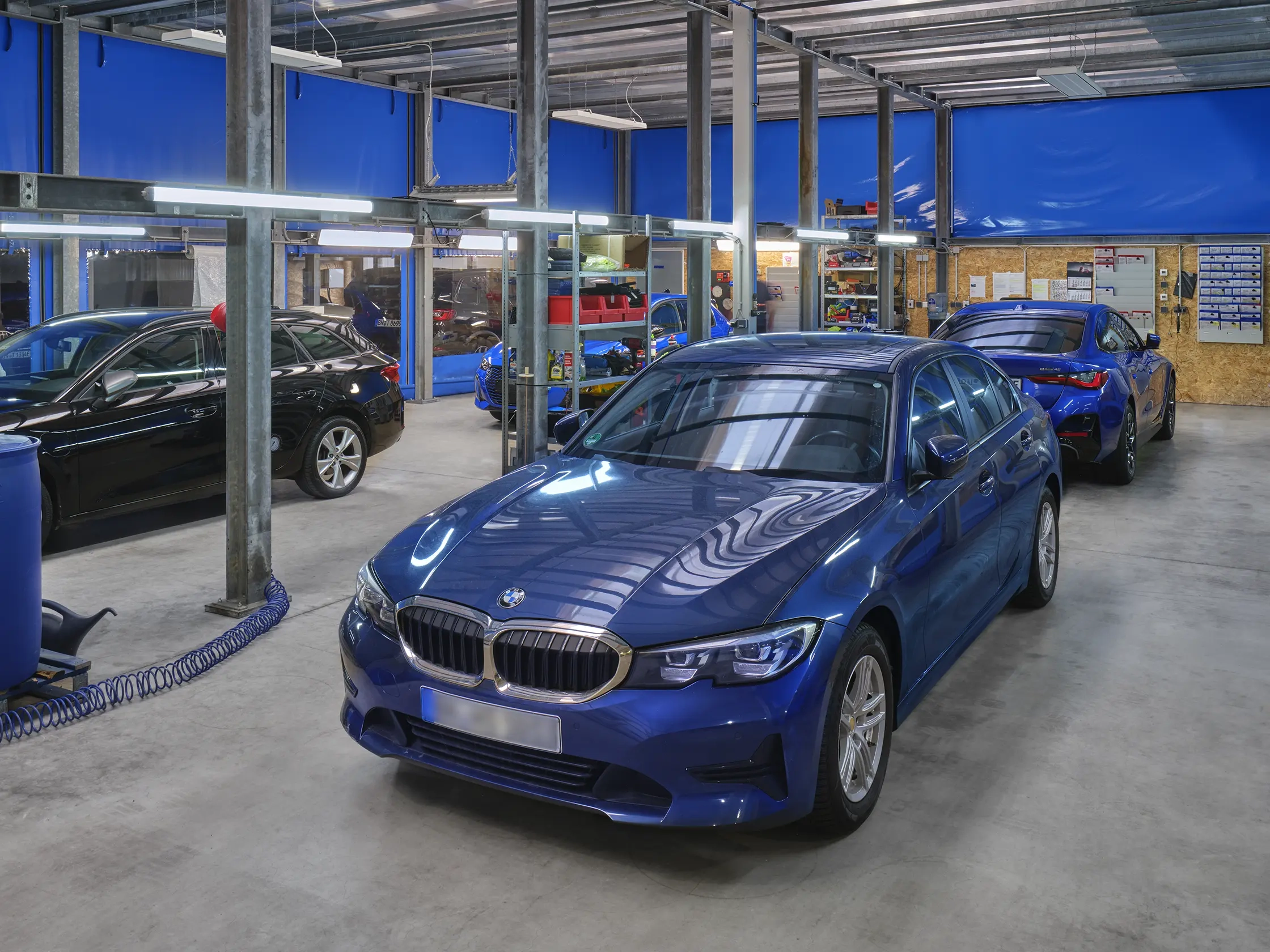 Eine Fahrzeugaufbereitungswerkstatt mit mehreren Autos. Im Vordergrund steht ein blauer BMW. Im Hintergrund sind weitere Fahrzeuge sichtbar, darunter ein schwarzes Auto und ein rotes Auto.