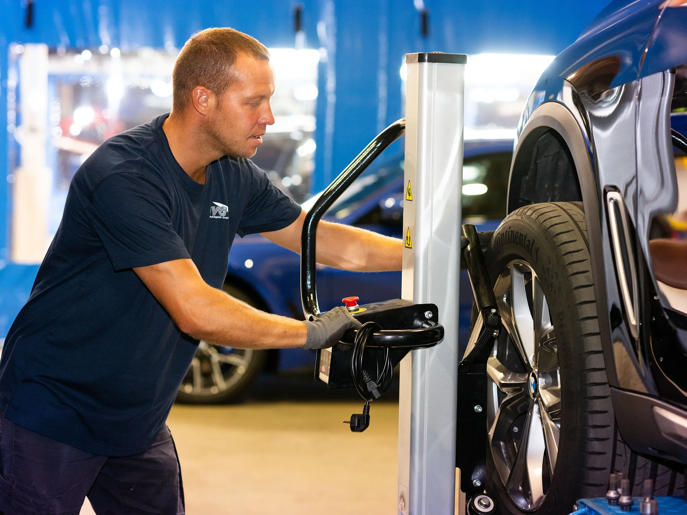 Ein Mitarbeiter von JVG wechselt einen Reifen an einem Fahrzeug. Er trägt ein dunkelblaues T-Shirt und Handschuhe. Im Hintergrund sind weitere Autos sichtbar.