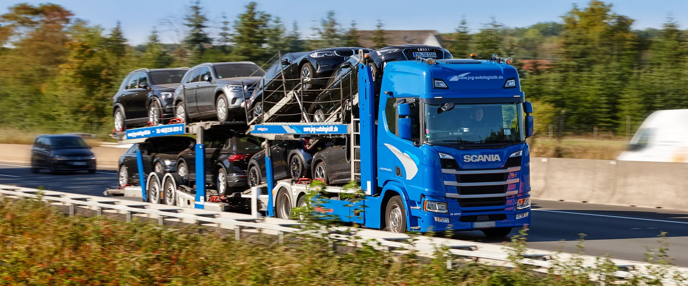 Blauer Scania Lkw von JVG transportiert mehrere schwarze Autos auf einem Anhänger auf der Autobahn. Im Hintergrund Bäume und eine andere Fahrbahn.