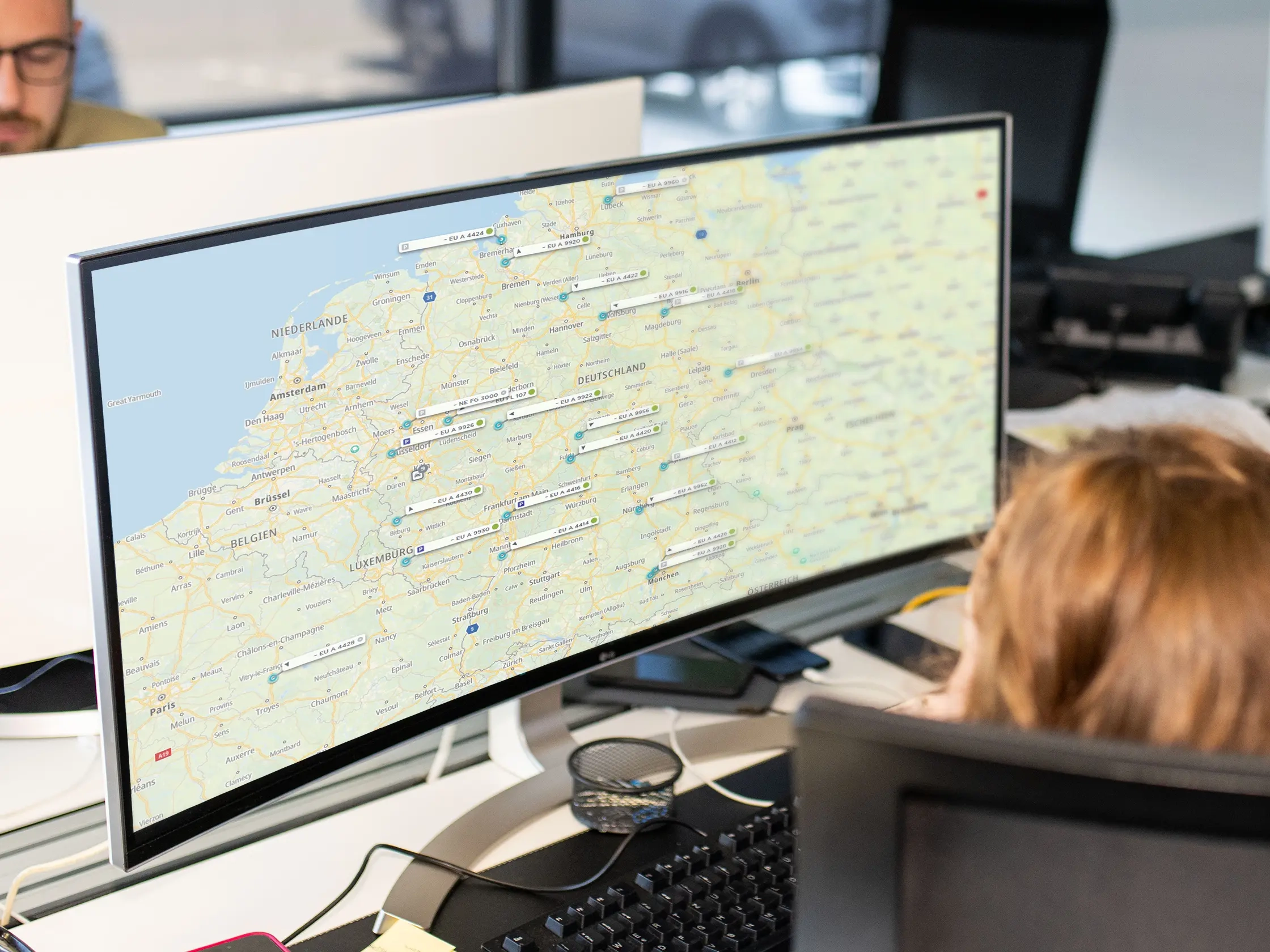 Eine Person sitzt vor einem großen gebogenen Monitor, der eine Karte mit verschiedenen Standorten in Europa zeigt. Auf dem Tisch sind Tastatur und ein Stift sichtbar.