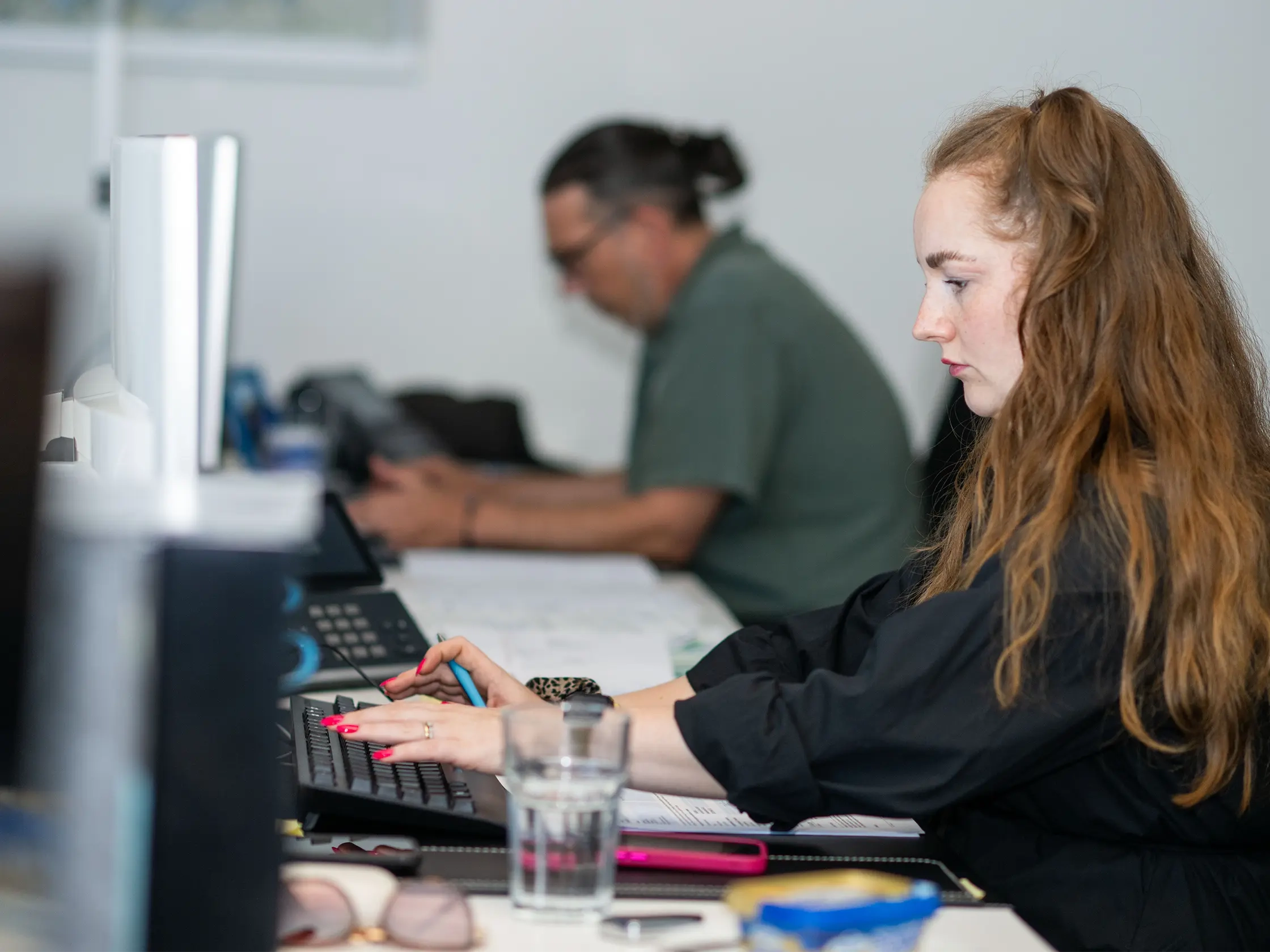 Eine Person mit langen, lockigen, braunen Haaren tippt an einer Tastatur. Im Hintergrund ist eine weitere Person am Arbeiten.