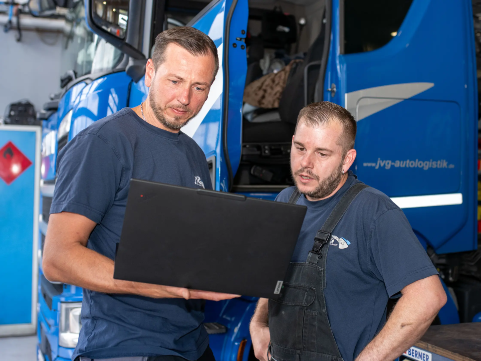 Zwei JVG Mitarbeiter in Arbeitskleidung, einer hält einen Laptop, der andere steht daneben. Im Hintergrund ein blauer Lkw.