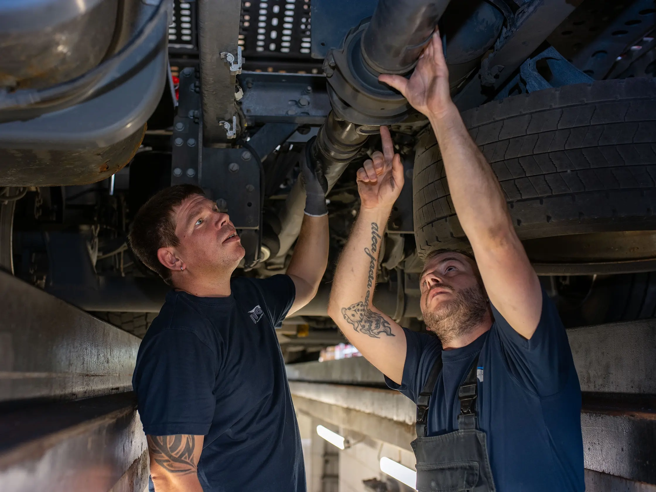 Zwei JVG-Mitarbeiter arbeiten unter einem LKW, einer zeigt auf ein Bauteil. Beide tragen Arbeitskleidung.